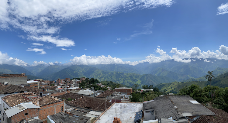 Panoramic view of a small town in Colombia, with terracotta rooftops in the foreground and lush green mountains rising under a blue sky with clouds